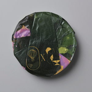 A round, wrapped product, likely a tea cake or puck, wrapped in dark green textured paper with gold foil designs and accents of purple and green, set against a solid gray background.