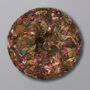 A round block of compressed tea leaves intertwined with pink rose petals on a gray background, creating a rustic and floral aesthetic.