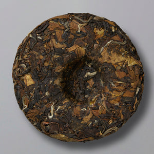 A round, tightly compressed cake of dark brown and tan tea leaves with a textured surface, set against a plain gray background.