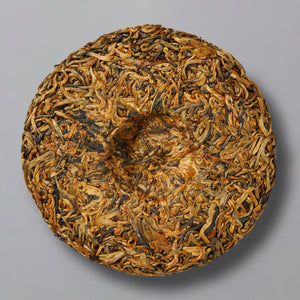 A round disc of tightly packed, dried golden bud black tea leaves, resembling a cake, sits on a plain gray background, conveying a sense of natural texture.