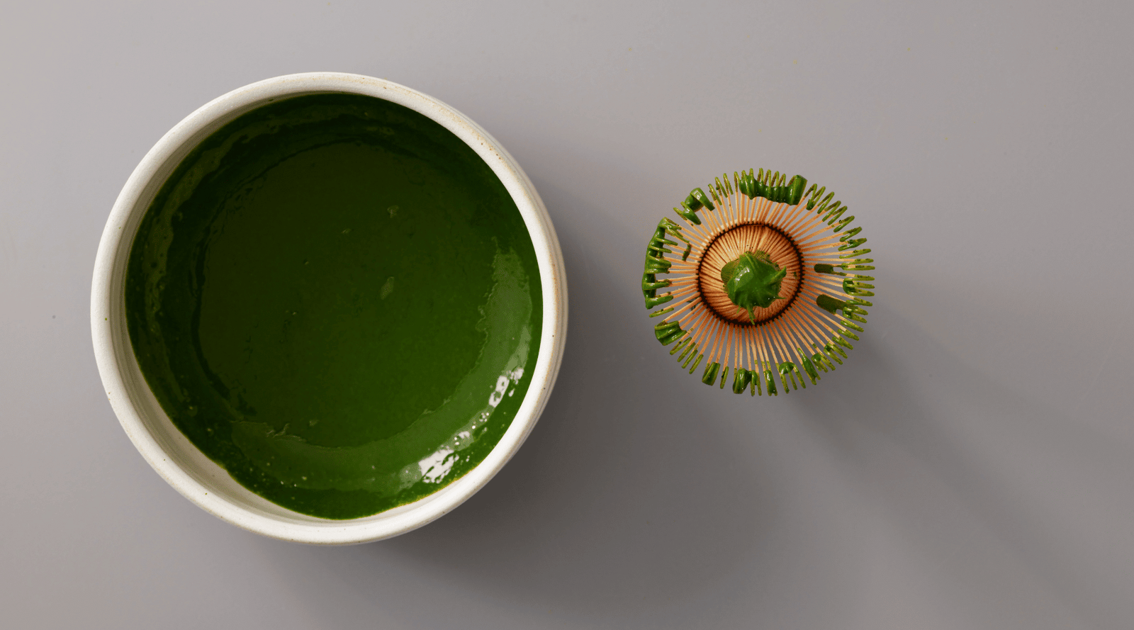A bowl of vibrant green matcha tea on a gray surface, with a bamboo whisk beside it. The setting conveys a minimalist, calming atmosphere.