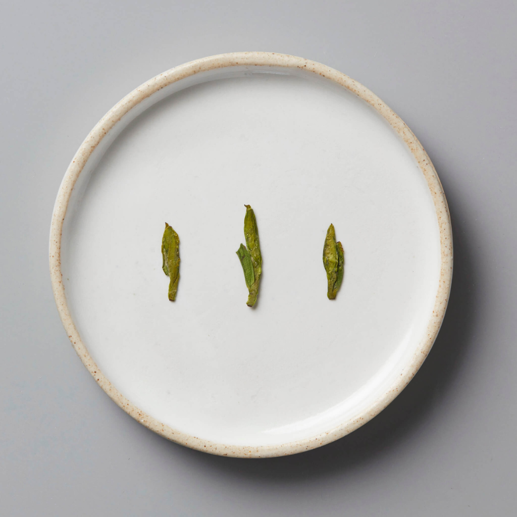 Three small green leaves are evenly spaced on a simple white ceramic plate against a gray background, conveying minimalism and elegance. Leaves are flat and dry indicating pan-firing.