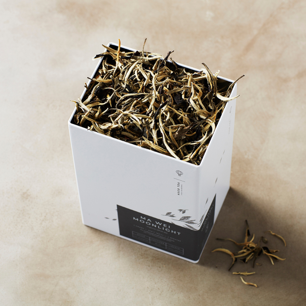 Open white box filled with dried tea leaves, labeled "Moonlight." Some leaves are scattered beside it on a beige surface, creating a rustic feel.