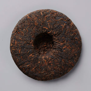 Circular compressed cake of DIGESTIF shou puer tea with dark brown and reddish tones and textured surface on gray background