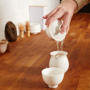 GAIWAN HUGO: Pouring tea from white porcelain gaiwan into serving vessel on wooden table with tea accessories