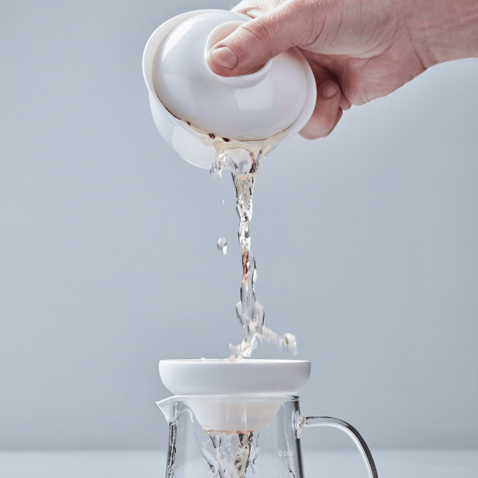 White porcelain filter pouring tea through strainer into glass pitcher, water streaming through perforated base
