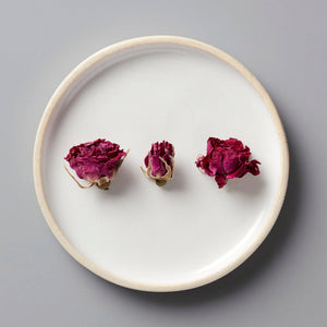 Three dried ROSE tisane buds arranged on a white ceramic plate with beige rim