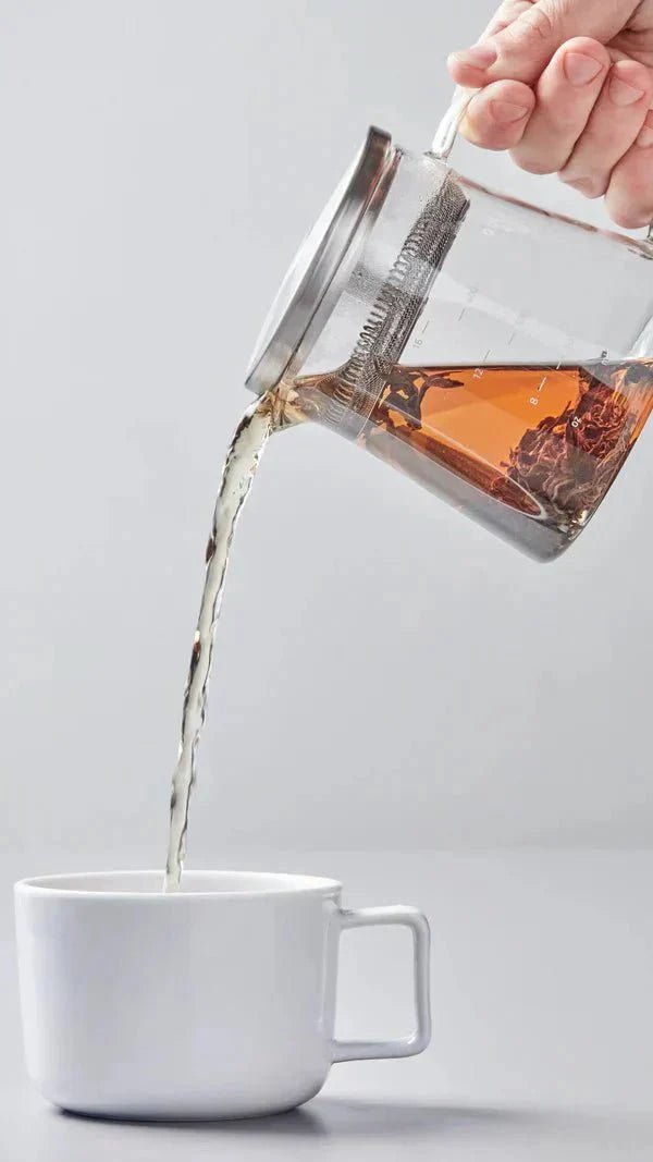 A person prepares tea in a traditional ceremony, pouring loose tea leaves from a bamboo scoop into a white gaiwan on a wooden tea tray. The setup includes white cups and a brown napkin, conveying a calm, ritualistic atmosphere.