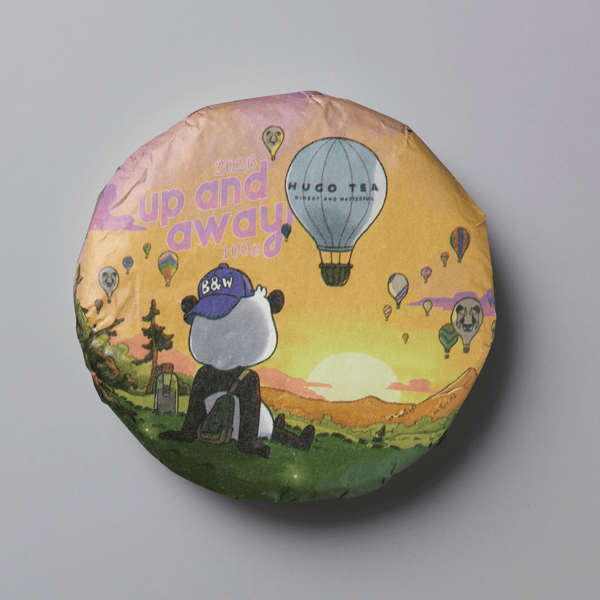 A round tea cake wrapped in illustrated paper packaging, featuring a panda sitting in a grassy field with hot air balloons and the text 'up and away' and 'HUGO TEA', photographed on a plain gray background.