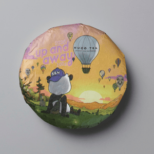 A round tea cake wrapped in illustrated paper packaging, featuring a panda sitting in a grassy field with hot air balloons and the text 'up and away' and 'HUGO TEA', photographed on a plain gray background.
