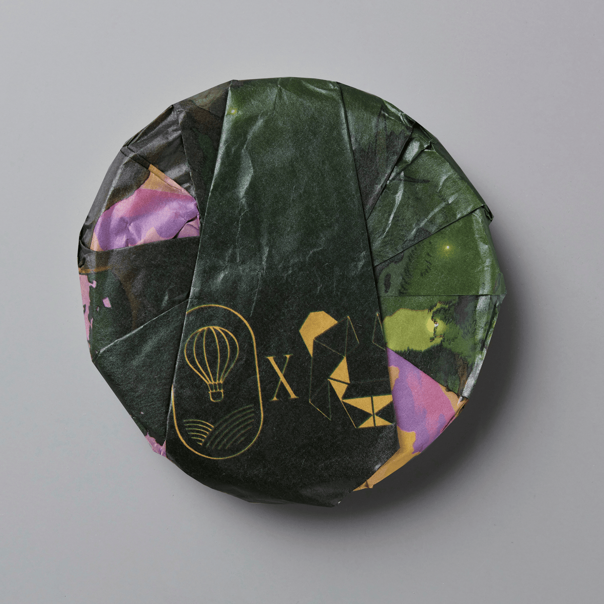 A round, wrapped product, likely a tea cake or puck, wrapped in dark green textured paper with gold foil designs and accents of purple and green, set against a solid gray background.