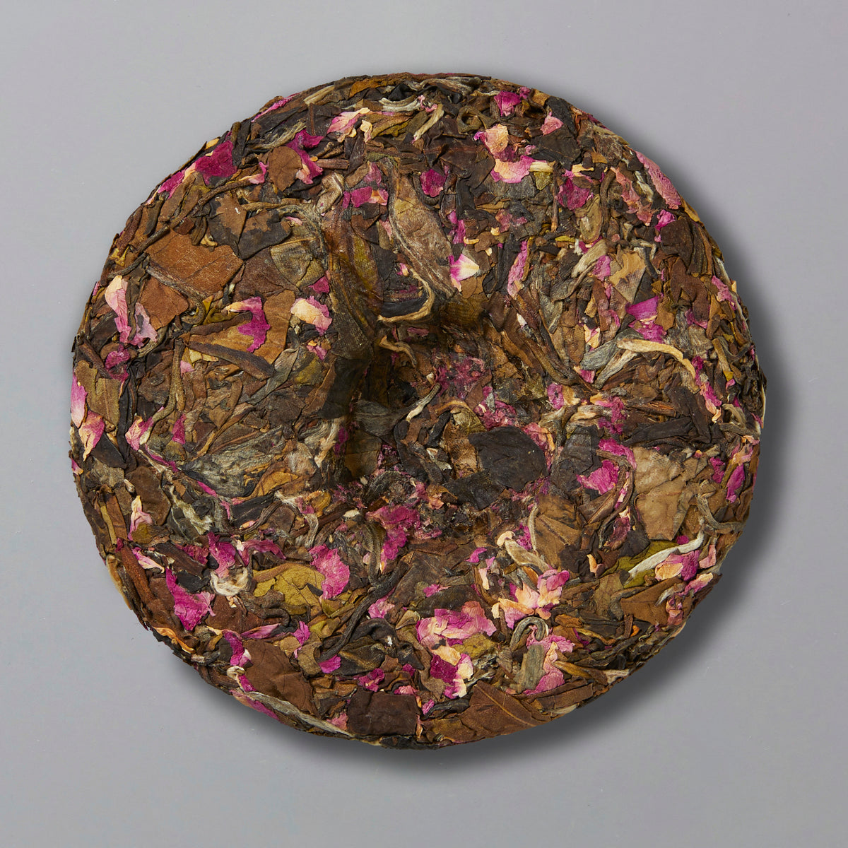 A round block of compressed tea leaves intertwined with pink rose petals on a gray background, creating a rustic and floral aesthetic.