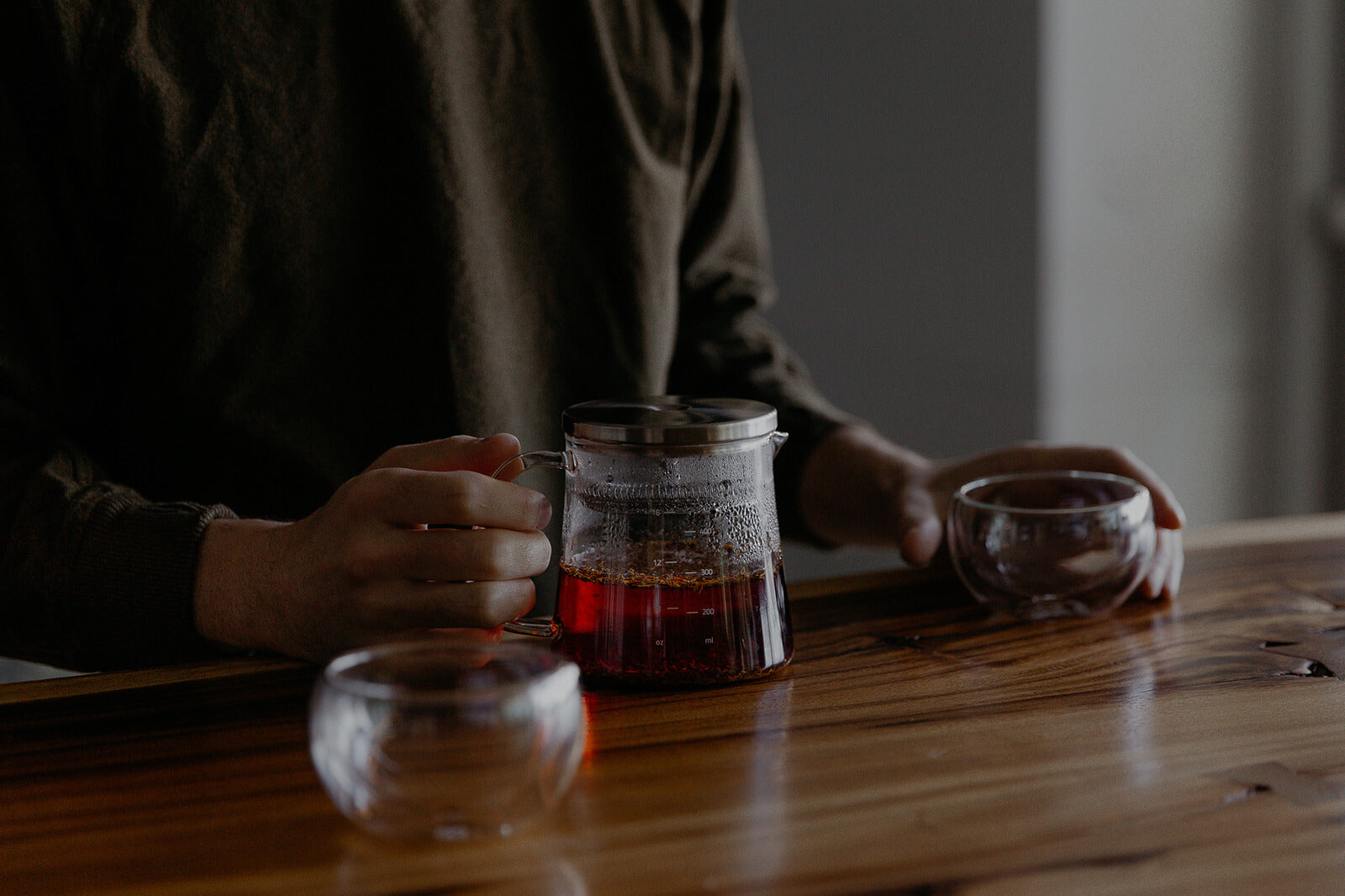 A person in a dark sweater holds a glass teapot with black tea, beside two empty glass cups on a wooden table, creating a cozy atmosphere.black 