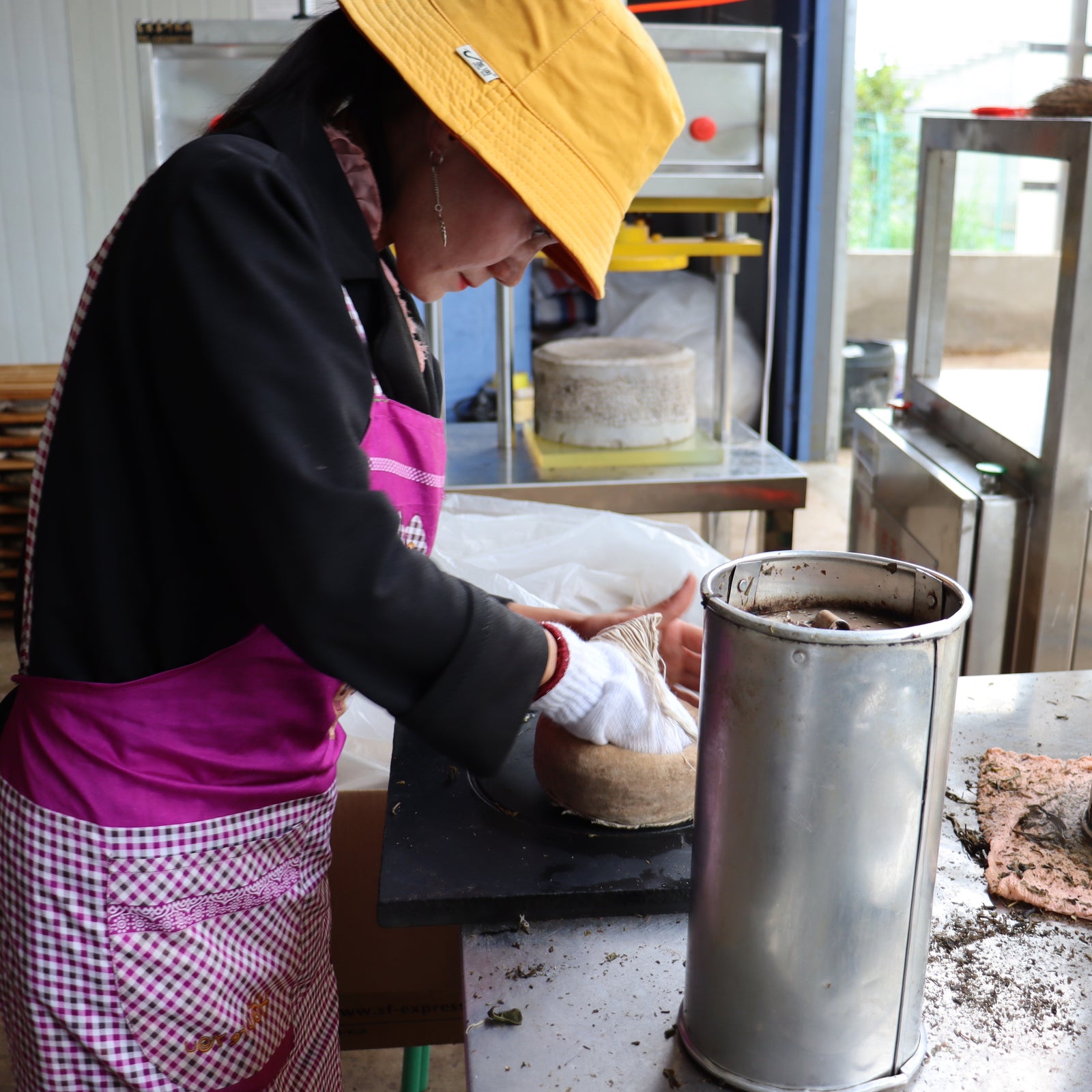 Woman in a yellow hat and apron presses tea leaves into a round mold at a workshop. She is focused, with various tools visible around her.
