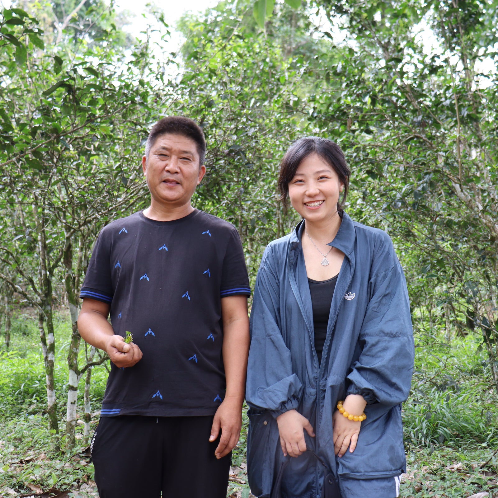 Two smiling individuals stand outdoors in a lush, green forest. The person on the left holds a small plant. Both wear casual clothing, conveying a happy and relaxed mood.