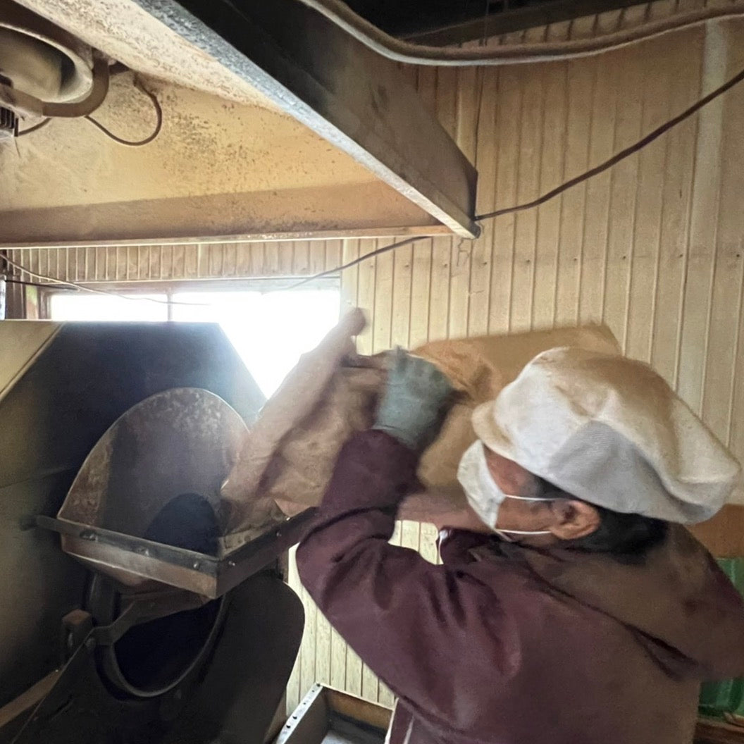 A worker in protective clothing and mask pours a large sack of material into an industrial machine. The setting is industrial, with wooden panel walls.