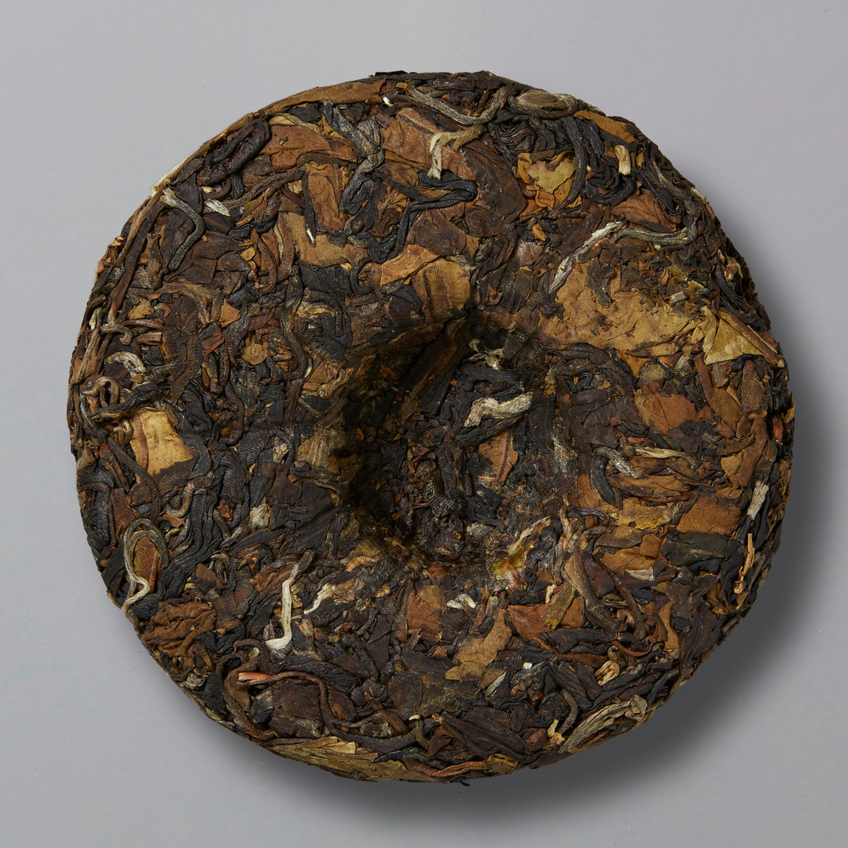 A round, tightly compressed cake of dark brown and tan tea leaves with a textured surface, set against a plain gray background.