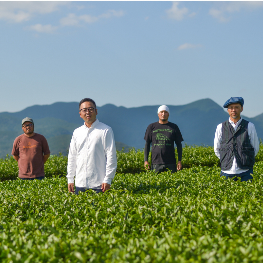 Four men stand in a lush green tea field under a clear blue sky, with mountains in the background. The mood is serene and peaceful.