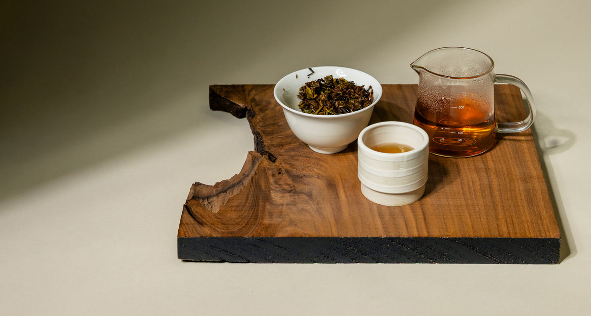 A wooden tea serving set on a beige surface, featuring a dark wood tray with a live edge, a white bowl of loose-leaf tea, a small white cup, and a glass pitcher filled with amber tea.