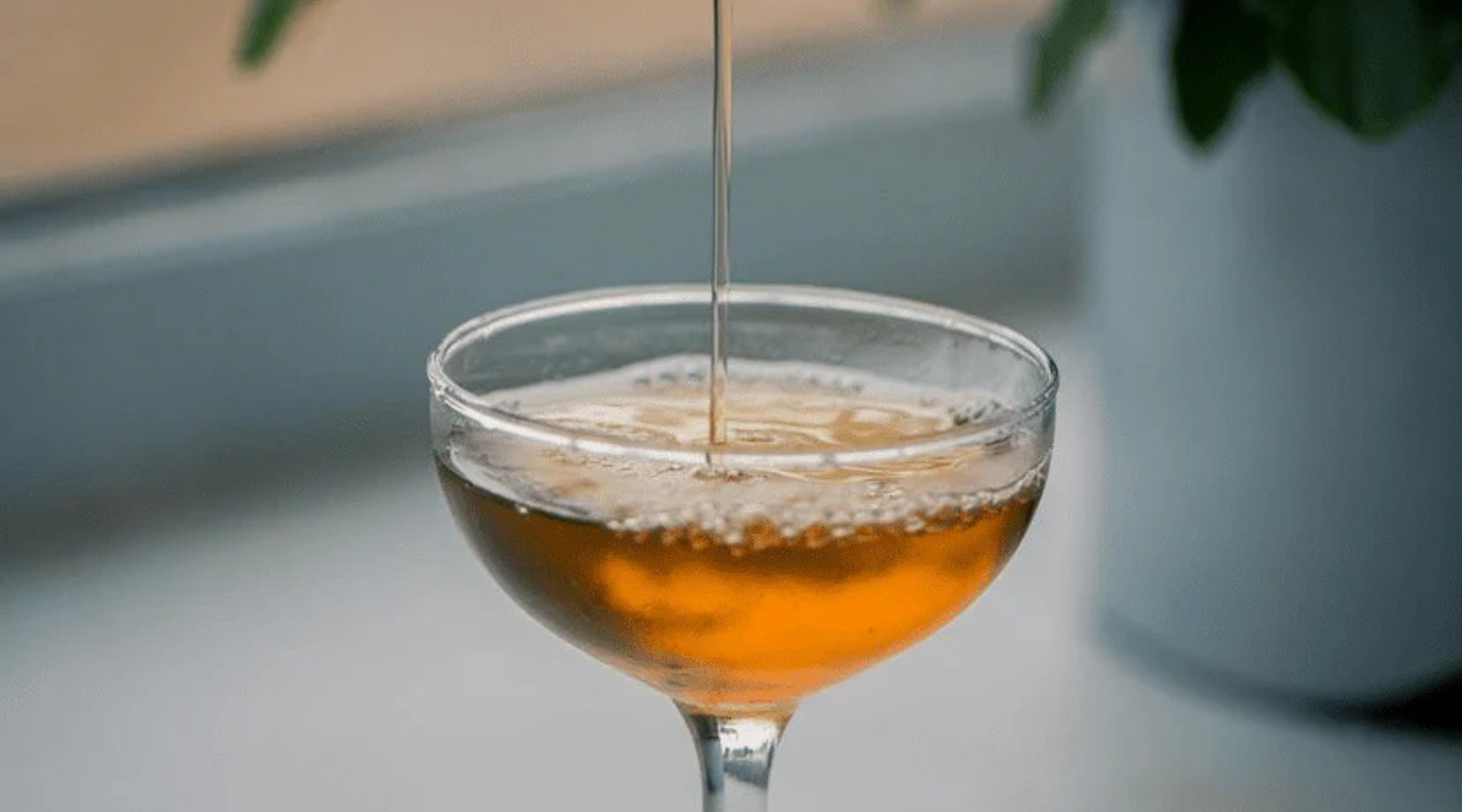 A stream of amber liquid is poured into a glass, creating bubbles. The setting is calm with a soft-focus plant in the background, evoking a relaxed atmosphere.