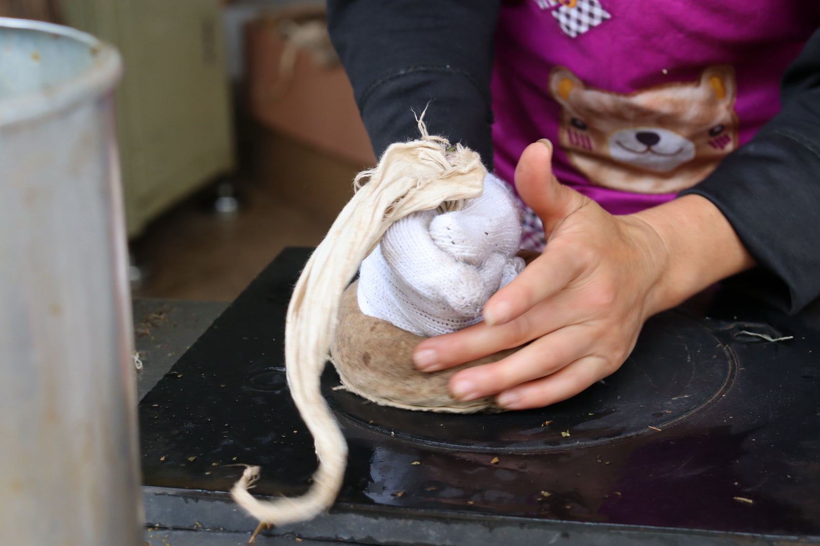 Hands firmly press a cloth-wrapped object against a dark surface. The steamed tea is packed into a cloth sack and shaped by hand. This is usually made into a discus called a "bing", or "cake".