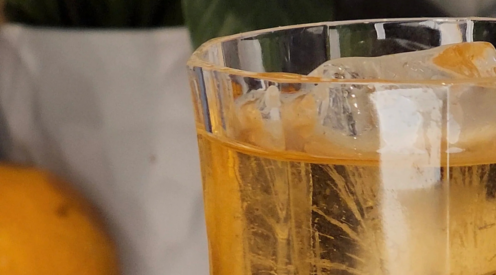 A close-up of a glass filled with amber-colored liquid and ice. A blurred lemon and greenery are in the background, creating a refreshing and vibrant tone.