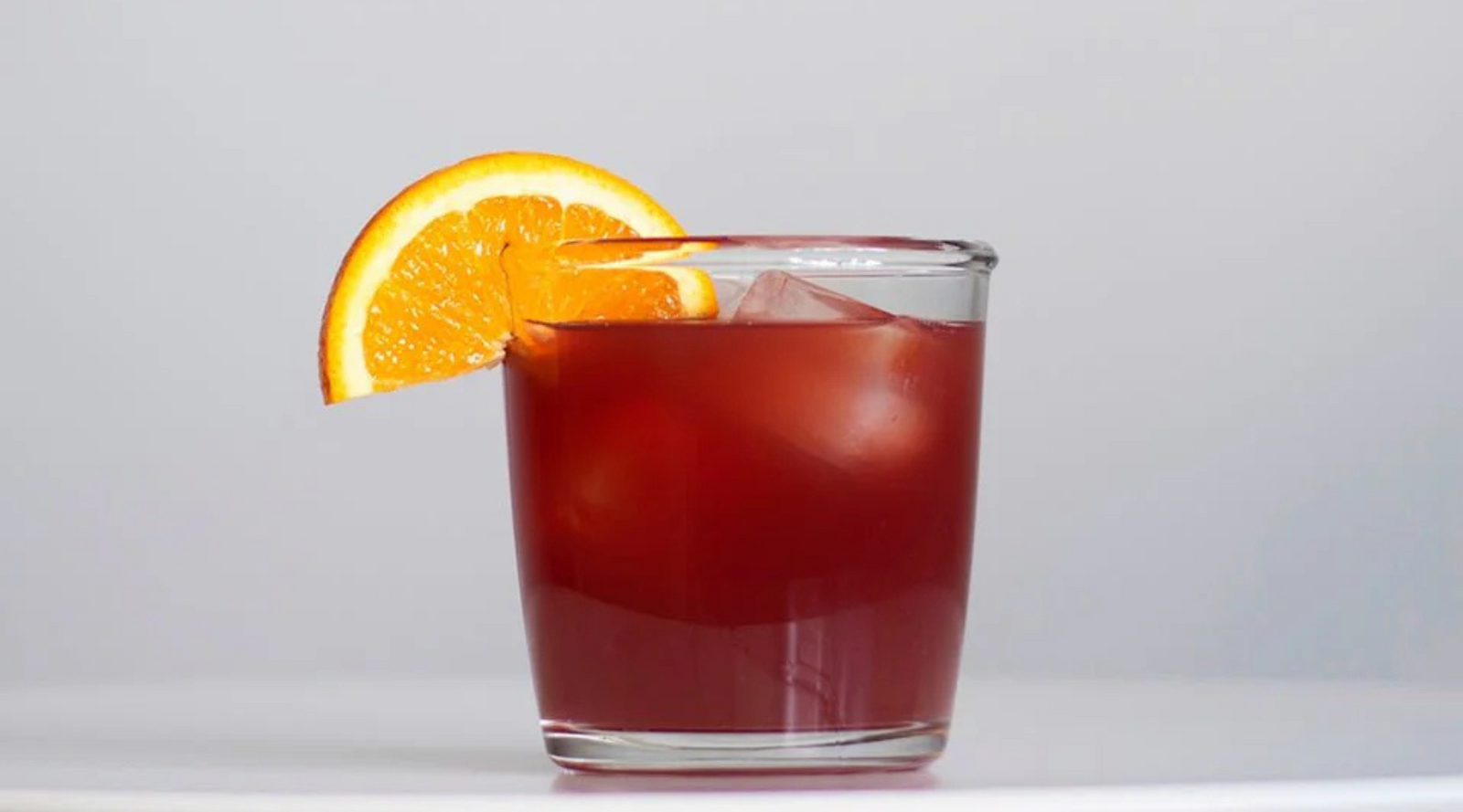 A glass of red iced sangria cocktail on a white surface, garnished with a slice of orange. The drink looks refreshing against a light gray background.