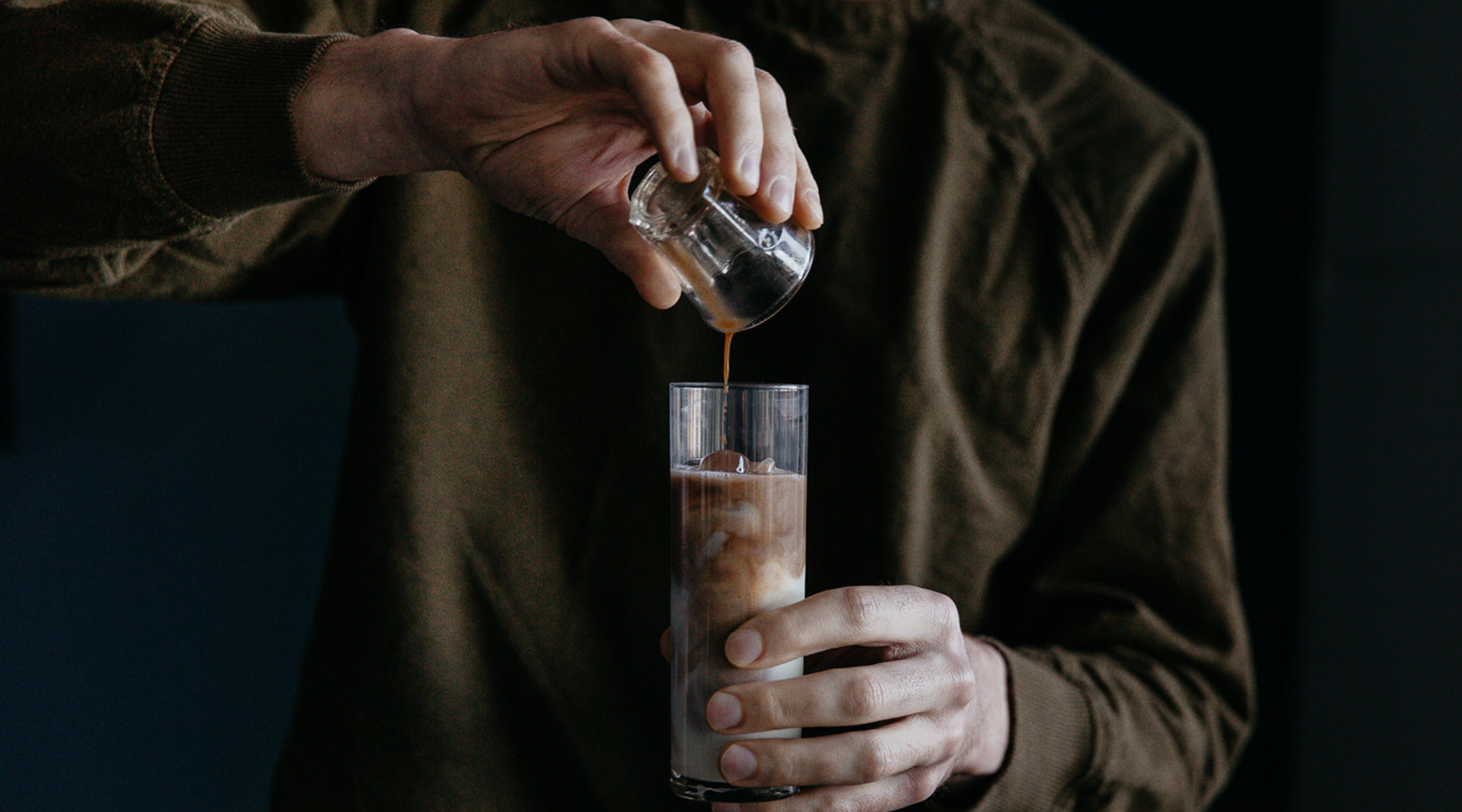 A person pours chai into a tall glass filled with ice and milk, creating a layered effect. The setting is dimly lit, conveying a calm and focused mood.