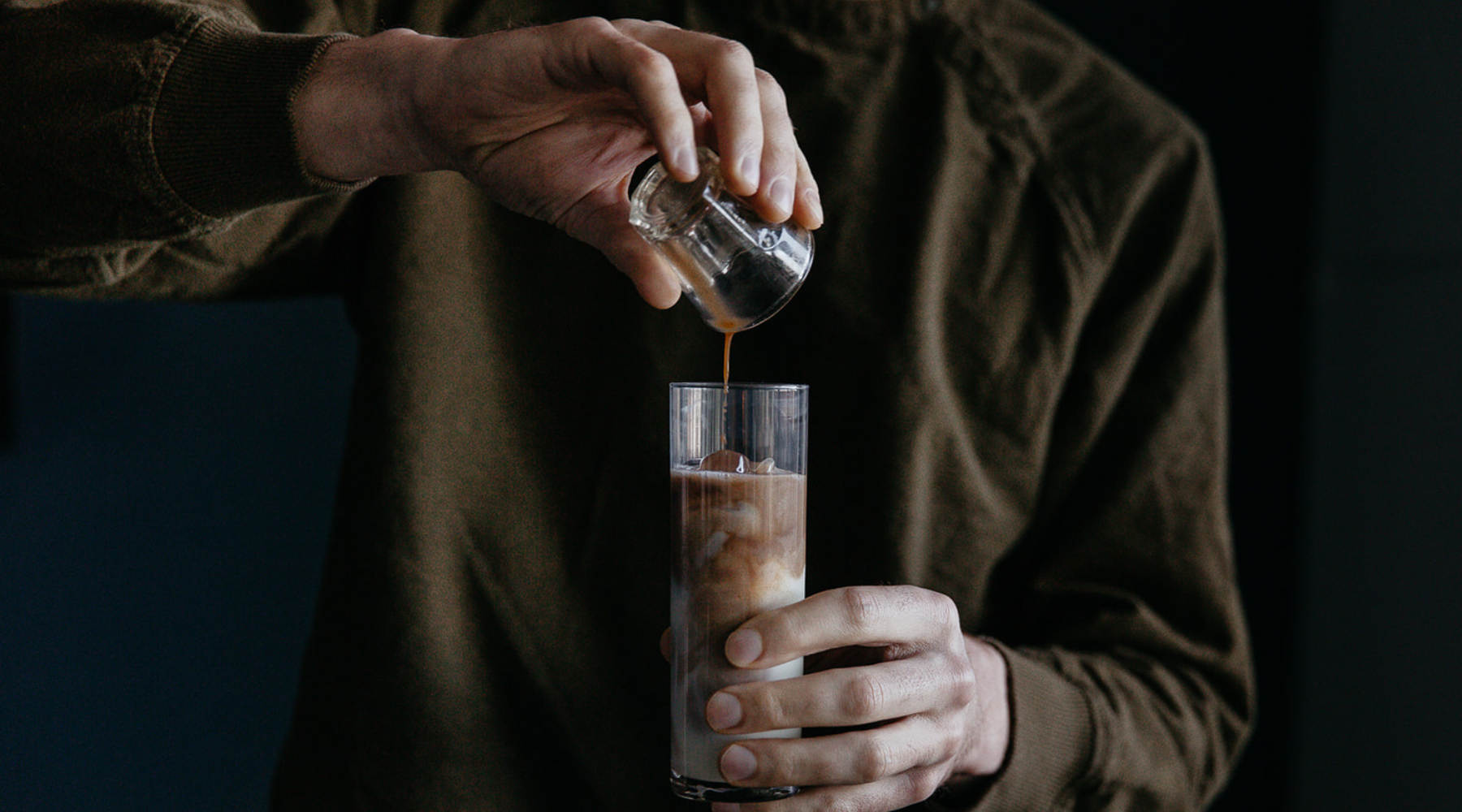 A person pours chai into a tall glass filled with ice and milk, creating a layered effect. The setting is dimly lit, conveying a calm and focused mood.