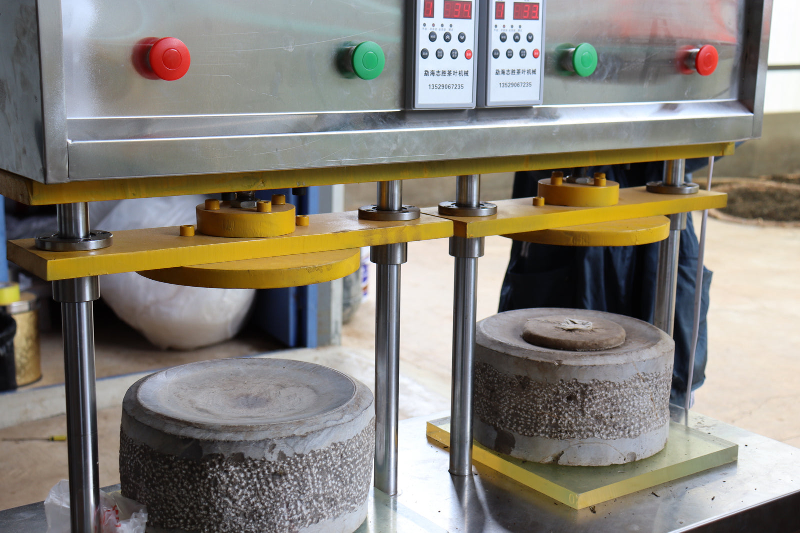 Industrial hydraulic machine pressing two concrete cylinders; displays show numbers, with red and green buttons. The setting is a workshop. 