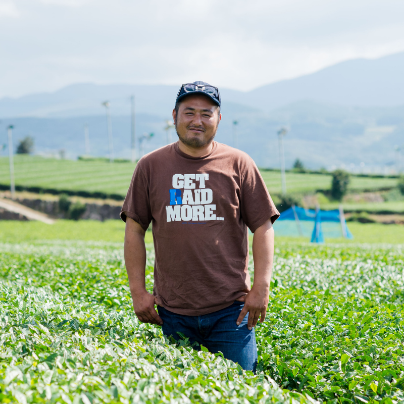 A smiling man in a brown T-shirt stands in a lush green field, set against a backdrop of rolling hills and a cloudy sky, conveying a serene and joyful mood.