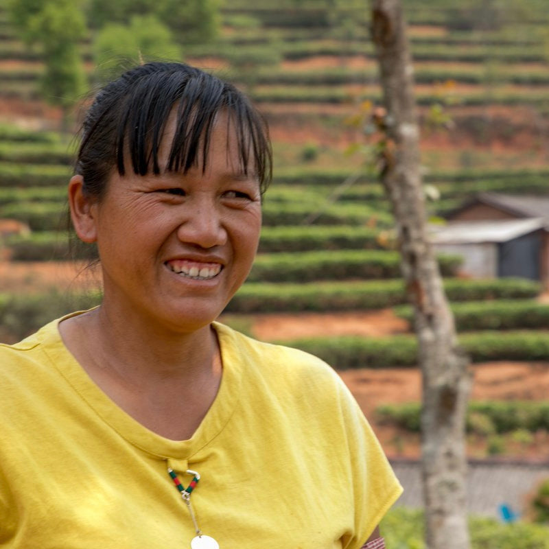 Smiling woman in a yellow shirt stands in front of lush, green terraced fields, conveying a serene and cheerful atmosphere in a rural setting.