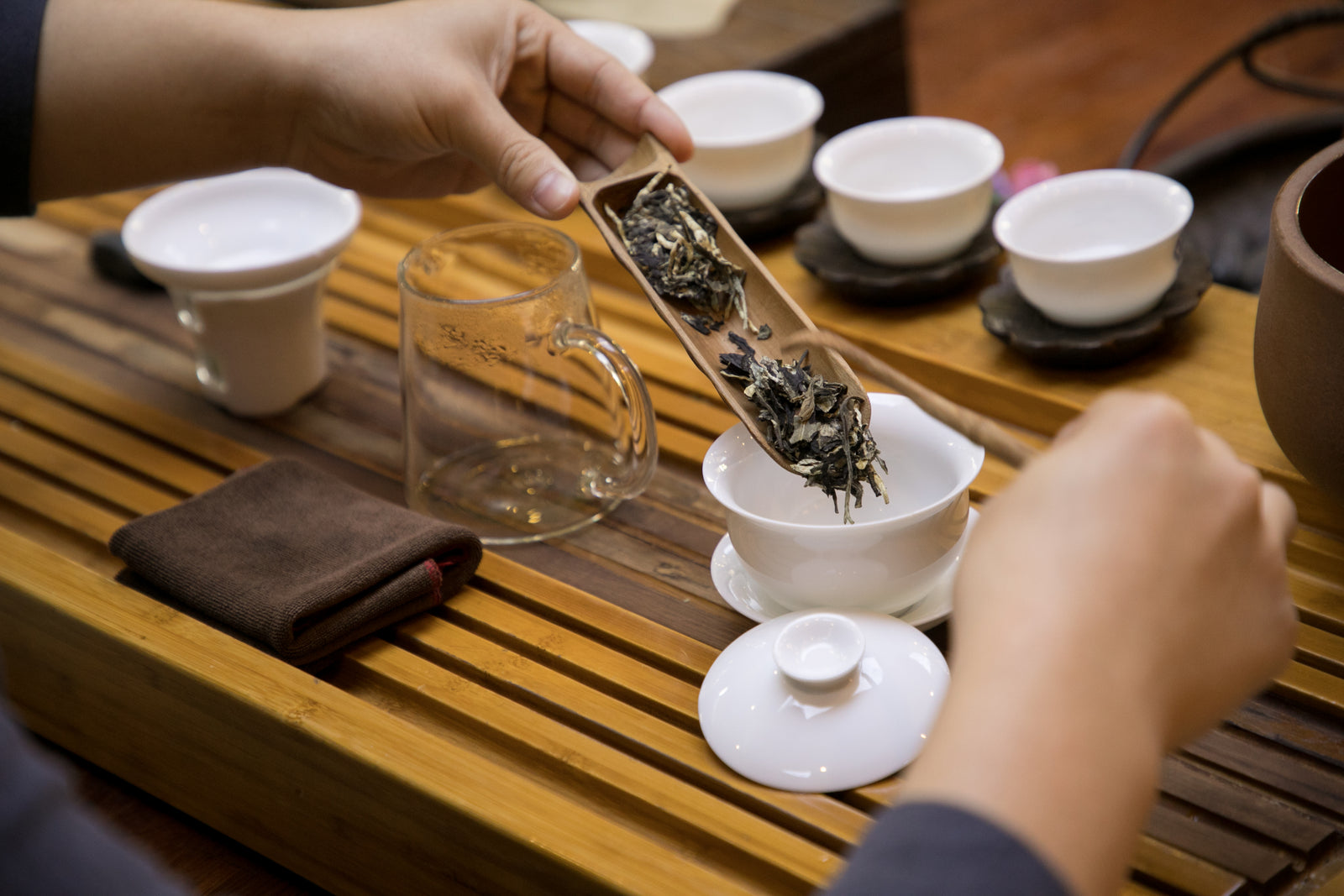 A person prepares tea in a traditional ceremony, pouring loose tea leaves from a bamboo scoop into a white gaiwan on a wooden tea tray. The setup includes white cups and a brown napkin, conveying a calm, ritualistic atmosphere.