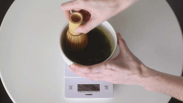Hands whisking matcha in a chawan using a bamboo chasen on a digital scale — demonstrating precise ratio and water measurement for brewing ceremonial matcha.