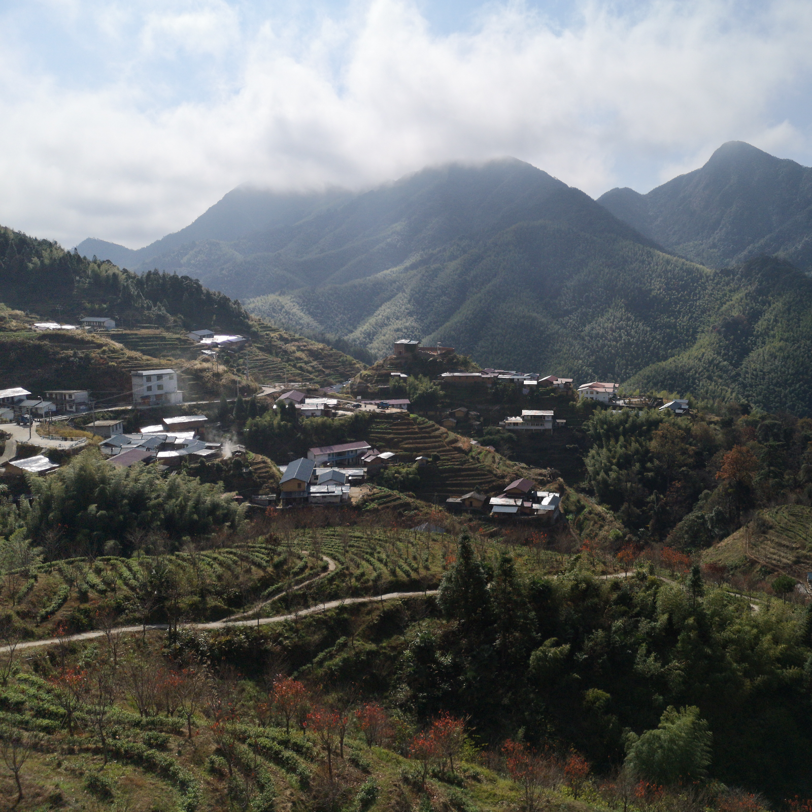 A scenic rural village nestled within lush terraced hills on a cloudy day. Mountain peaks loom in the background. The scene feels peaceful and serene.