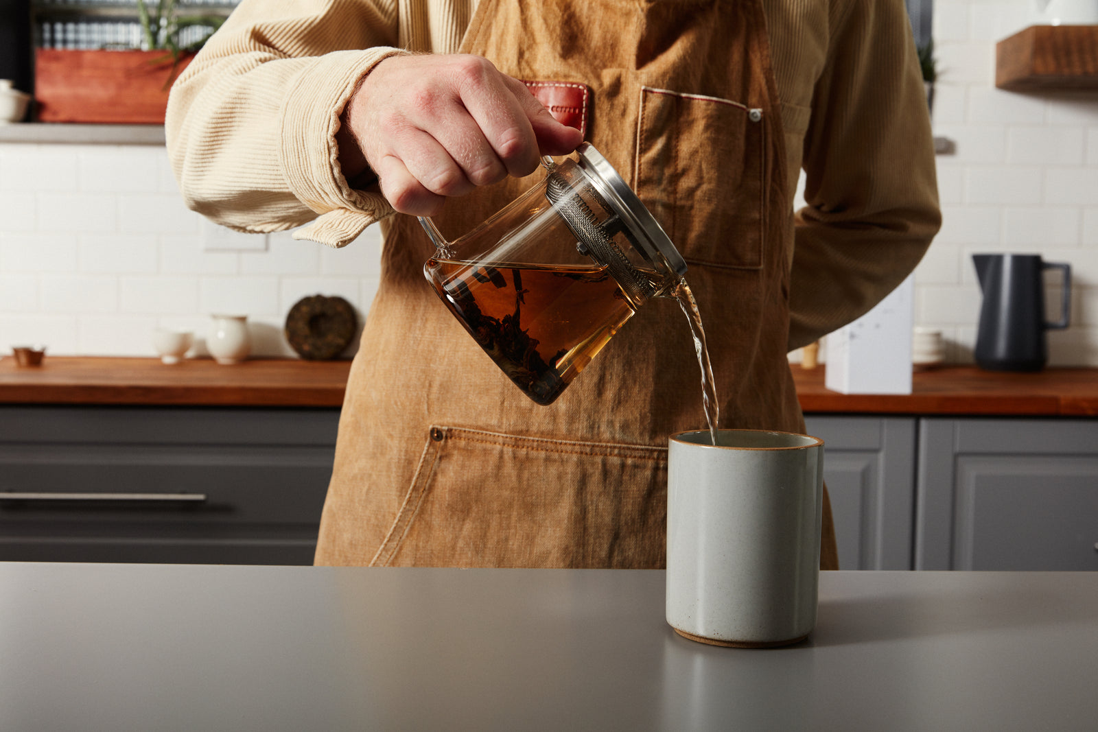 Person in a brown apron pouring tea from a glass pot into a gray mug on a kitchen counter. The background features white tiles and wooden accents.