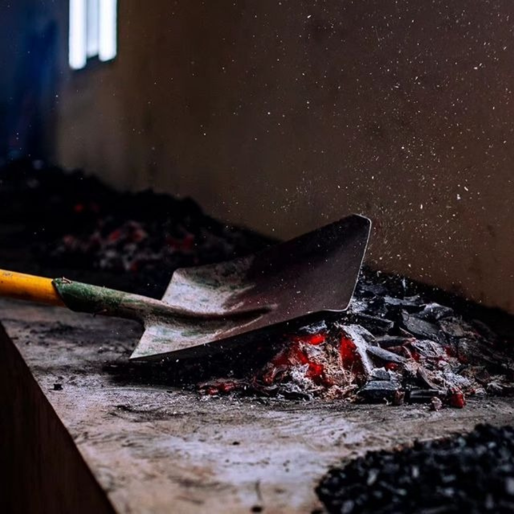 A shovel with a yellow handle moves through hot coals and ashes, sending sparks upward. The dim lighting and scattered debris convey a gritty atmosphere.