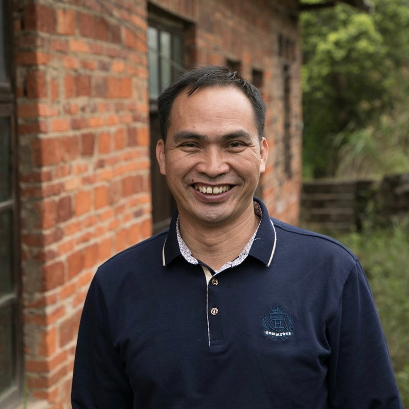 Smiling man in a navy polo shirt stands outdoors next to a rustic brick building and greenery, conveying a warm and welcoming atmosphere.