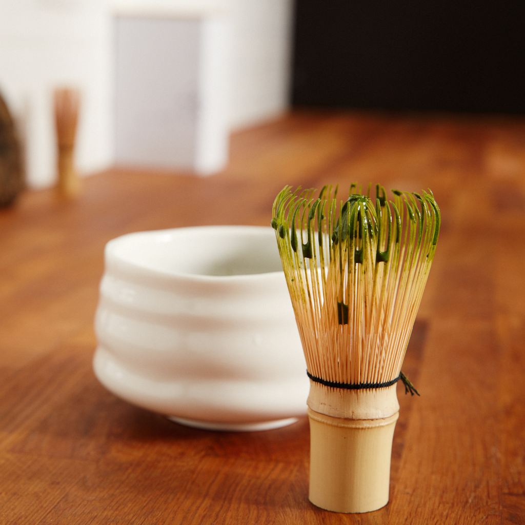 Bamboo chasen whisk coated in freshly prepared matcha standing beside a white chawan tea bowl on a wooden table — traditional tools used for brewing ceremonial matcha.
