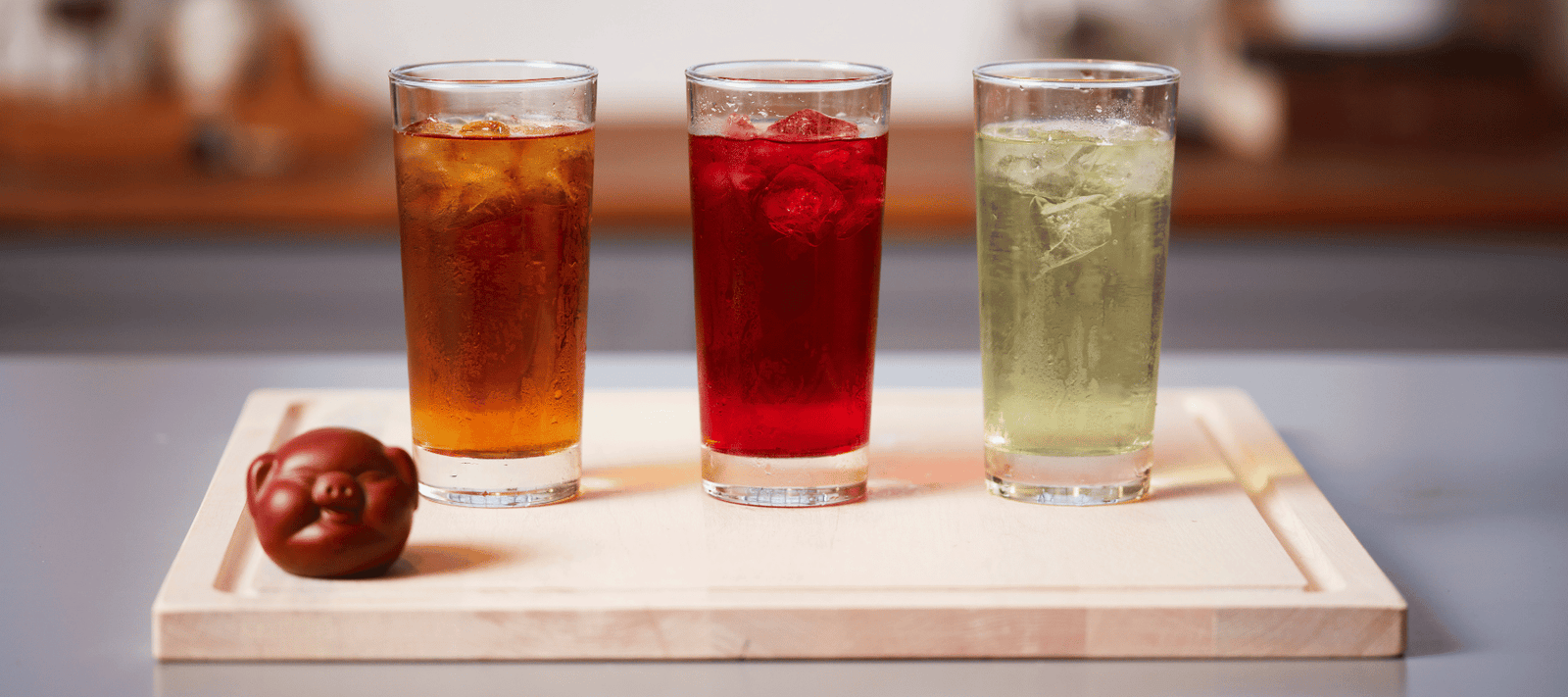 Three glasses of iced tea—amber, red, and pale green—sit on a wooden tray. A small, decorative brown object is placed on the tray beside the glasses.