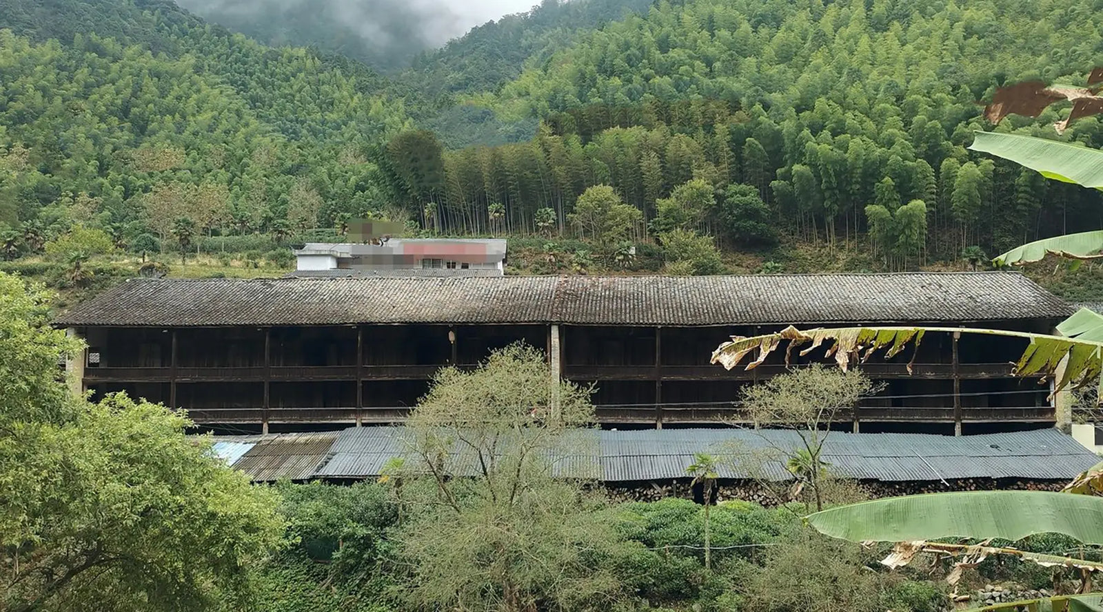 A rustic, multi-level wooden barn stands amidst lush green forests and fog-topped mountains, conveying a serene and tranquil rural landscape.