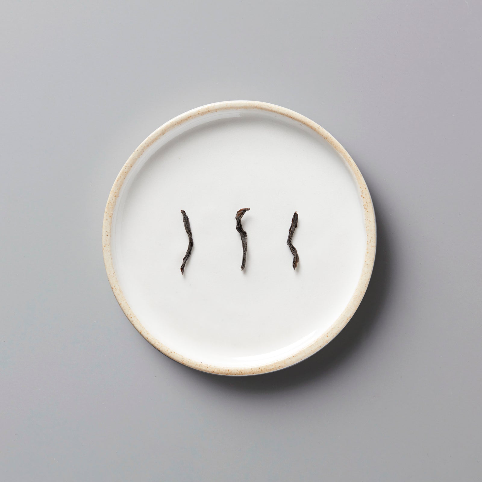 Three dark tea leaves of LINS RED black tea displayed on a round white ceramic plate with beige rim
