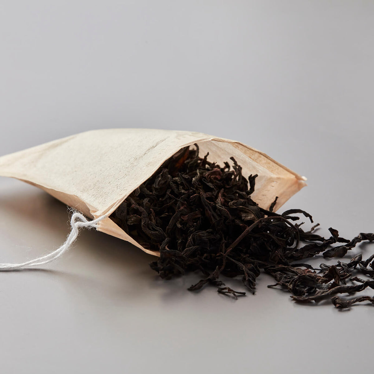 Drawstring sachets with loose black tea leaves spilling from unbleached cotton tea bag on gray background