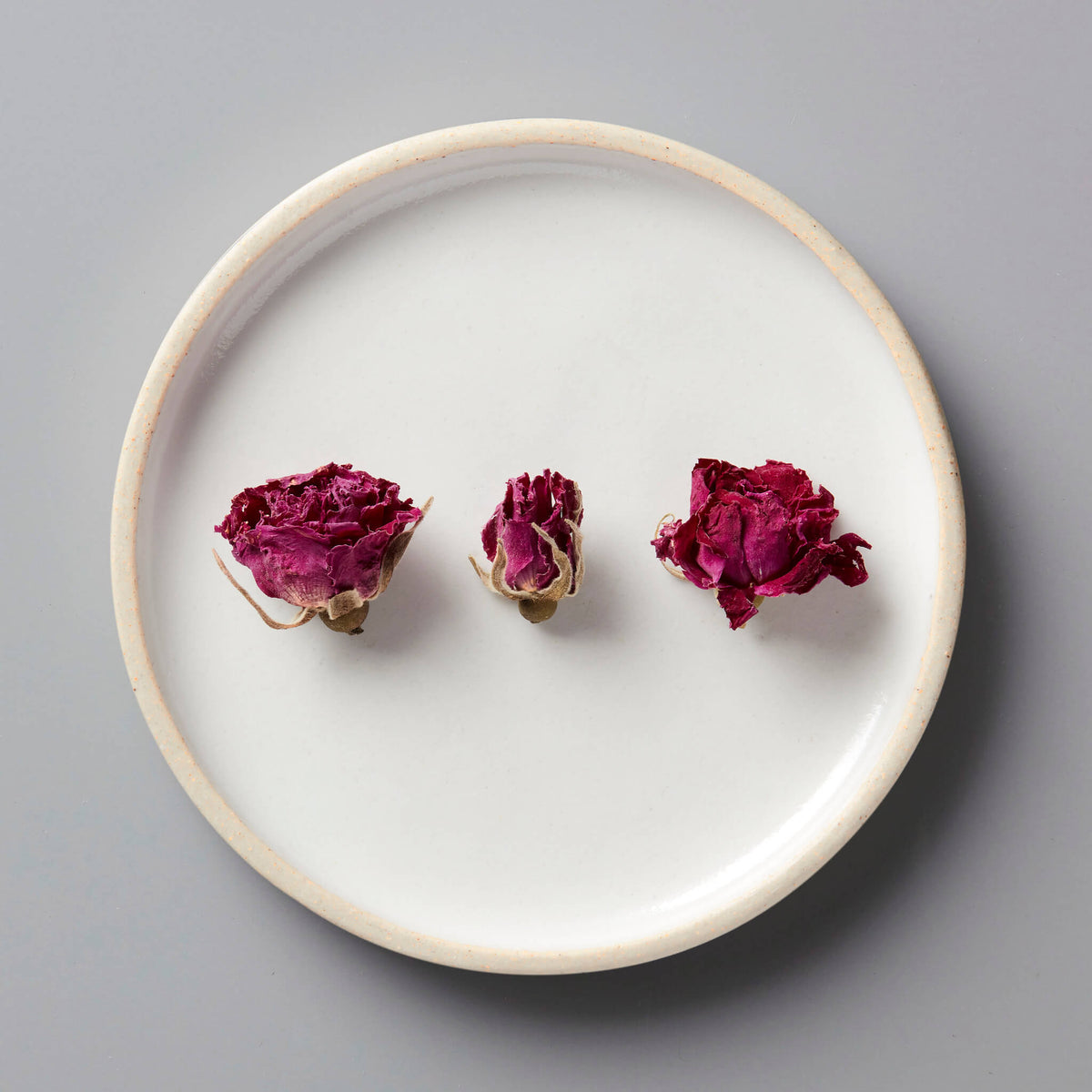 Three dried ROSE tisane buds arranged on a white ceramic plate with beige rim